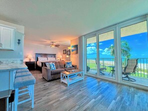 A spacious living area with a balcony overlooking a beach, a dining area with bar stools, and a bedroom with a large bed and a ceiling fan.