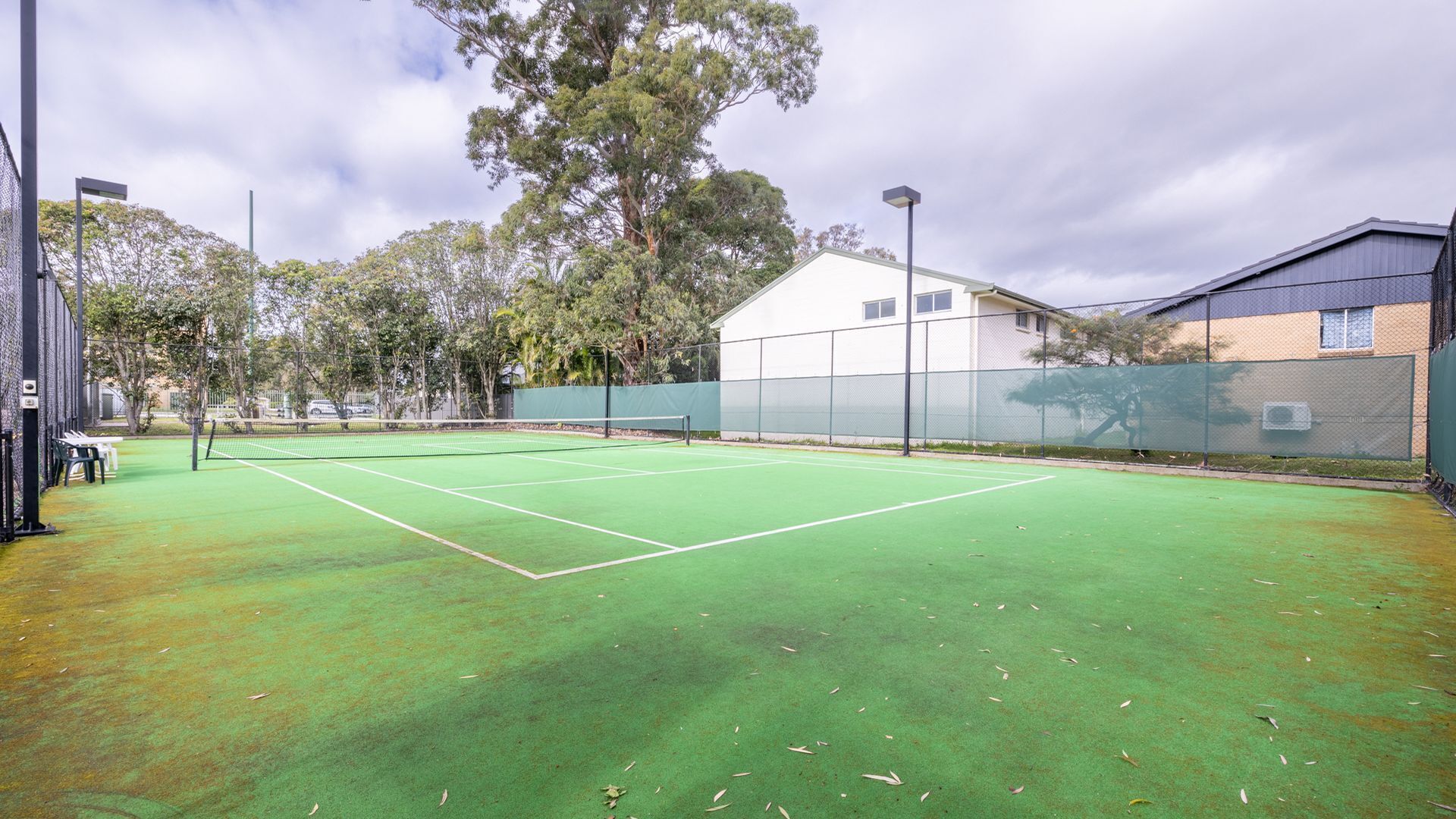 Un court de tennis en plein air, doté d’une surface verte, entouré d’arbres et bordé par un bâtiment.