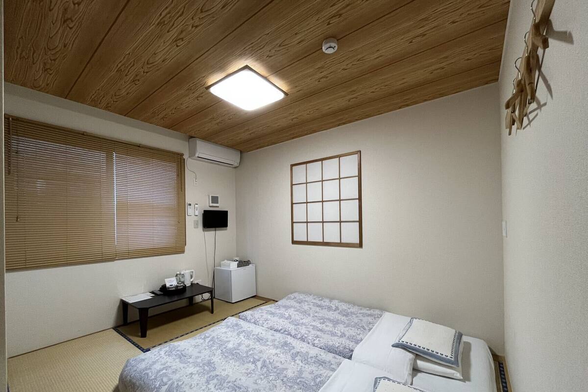 A room with a wooden ceiling, a bed with patterned bedding, a small table with a kettle, and a wall-mounted air conditioner.