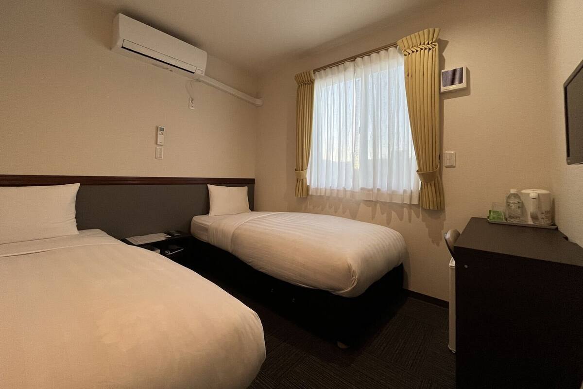 A hotel room with two beds, an air conditioning unit, a window with curtains, a desk with a telephone, and a mirror.