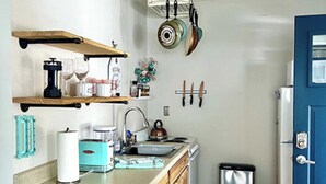 Une cuisine compacte avec des meubles en bois, un évier, une cuisinière et une poubelle. On y trouve des casseroles suspendues et une petite étagère contenant des ustensiles de cuisine.