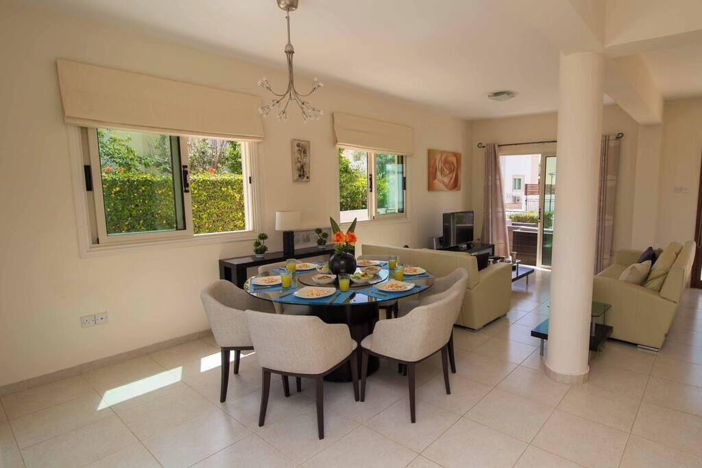A spacious living area with a round dining table, multiple chairs, a sofa, and a TV. Large windows provide natural light and a view of greenery outside.