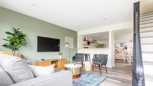 A modern living room with a grey sofa, a round coffee table, and two armchairs. A flat-screen TV is mounted on the wall, and there is a kitchen area visible in the background.