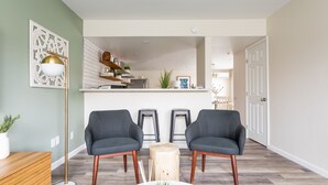 A modern living room with two grey armchairs, a wooden coffee table, and a floor lamp. A kitchen is visible in the background.