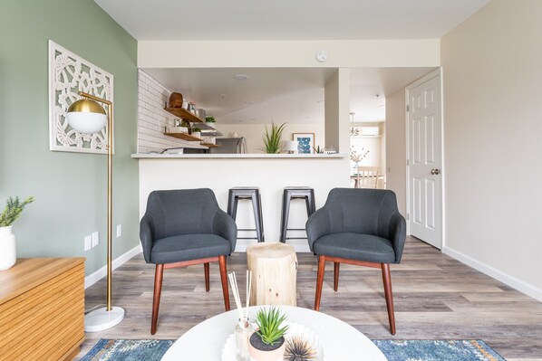 Una sala de estar moderna con dos sillones grises, una mesa de centro de madera y una lámpara de pie. Se observa una cocina al fondo.
