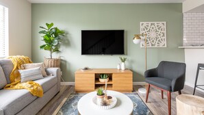 A living room with a sofa, a coffee table with decorative items, a TV mounted on the wall, and a side table with a lamp.
