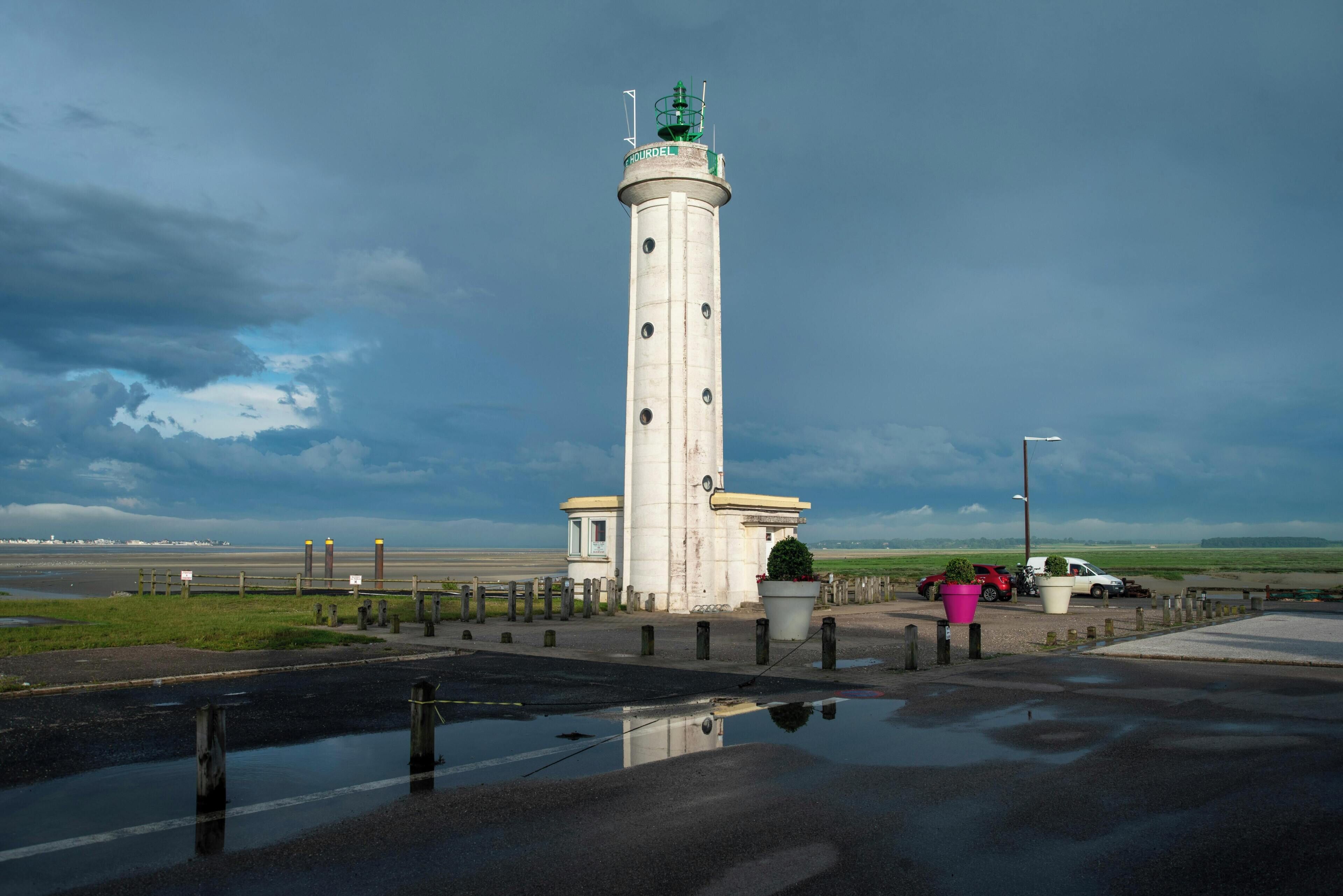 A tall white lighthouse with a green top, situated next to a road with puddles and parked cars.