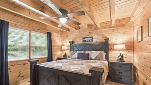 Een slaapkamer in een houten hut met een bed, nachtkastjes, een plafondventilator en een raam met jaloezieën.