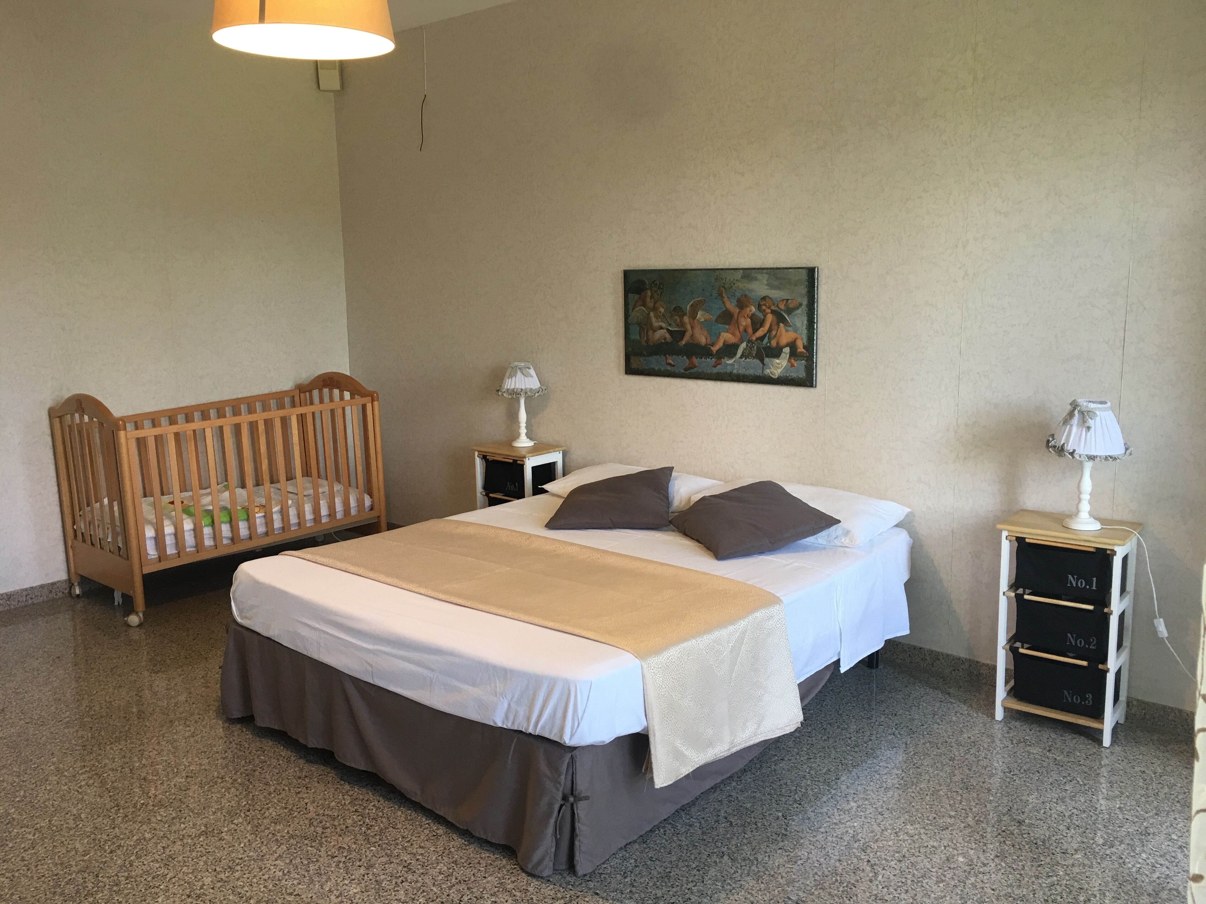 A bedroom with a large bed, a wooden crib, two bedside tables with lamps, and a painting on the wall.