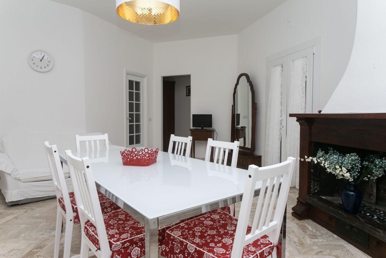 A dining area with a white table and chairs, a clock on the wall, and a fireplace with flowers.