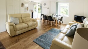A spacious living room with a beige sofa, a dining area with a table set for four, and a television on a wooden stand.