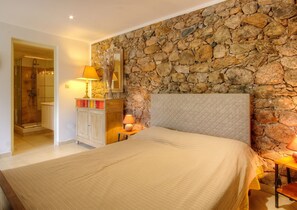 Une chambre avec un mur en pierre, un lit doté d’une tête de lit matelassée, une table de chevet avec une lampe et une commode avec un miroir.