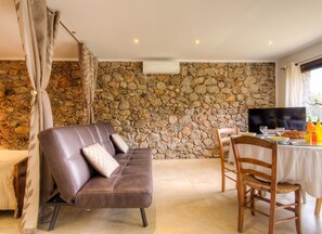 Habitació amb paret de pedra, un sofà, televisió i zona de menjador amb cadires i taula.
