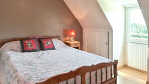 A bedroom with a sloped ceiling, a bed with patterned bedding, two decorative pillows, a bedside lamp, and a window with a view of greenery. - Homerez - House in Saint-Alban (Saint-Alban)