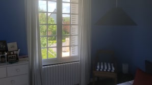 Quarto com uma janela grande, um radiador, uma cama, uma cadeira e uma mesinha de cabeceira.