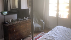 Quarto com cômoda de madeira, televisão, espelho, cama com colcha florida e janela com cortinas.