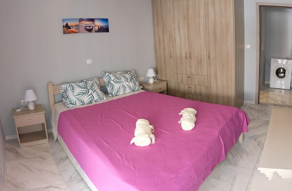 A bedroom with a large bed, a wooden wardrobe, a nightstand with a lamp, and a washing machine in the corner.