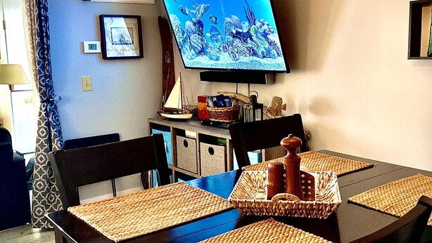 Een eethoek met een tafel voor vier personen, een aan de muur bevestigde televisie en een plafondlamp.