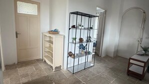Una habitación con piso de baldosas, una puerta blanca con un ventanal decorativo, un armario blanco, una estantería metálica negra y una pequeña mesa con una lámpara.