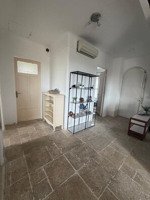 Une pièce avec un sol carrelé, une porte blanche ornée d’un vitrail, un meuble blanc, une étagère métallique noire et une petite table avec une lampe.