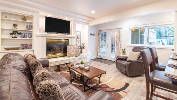 A spacious living room with a fireplace, a large flat-screen TV, a leather sofa, a dining area with a table and chairs, and a bookshelf filled with books and decorative items.