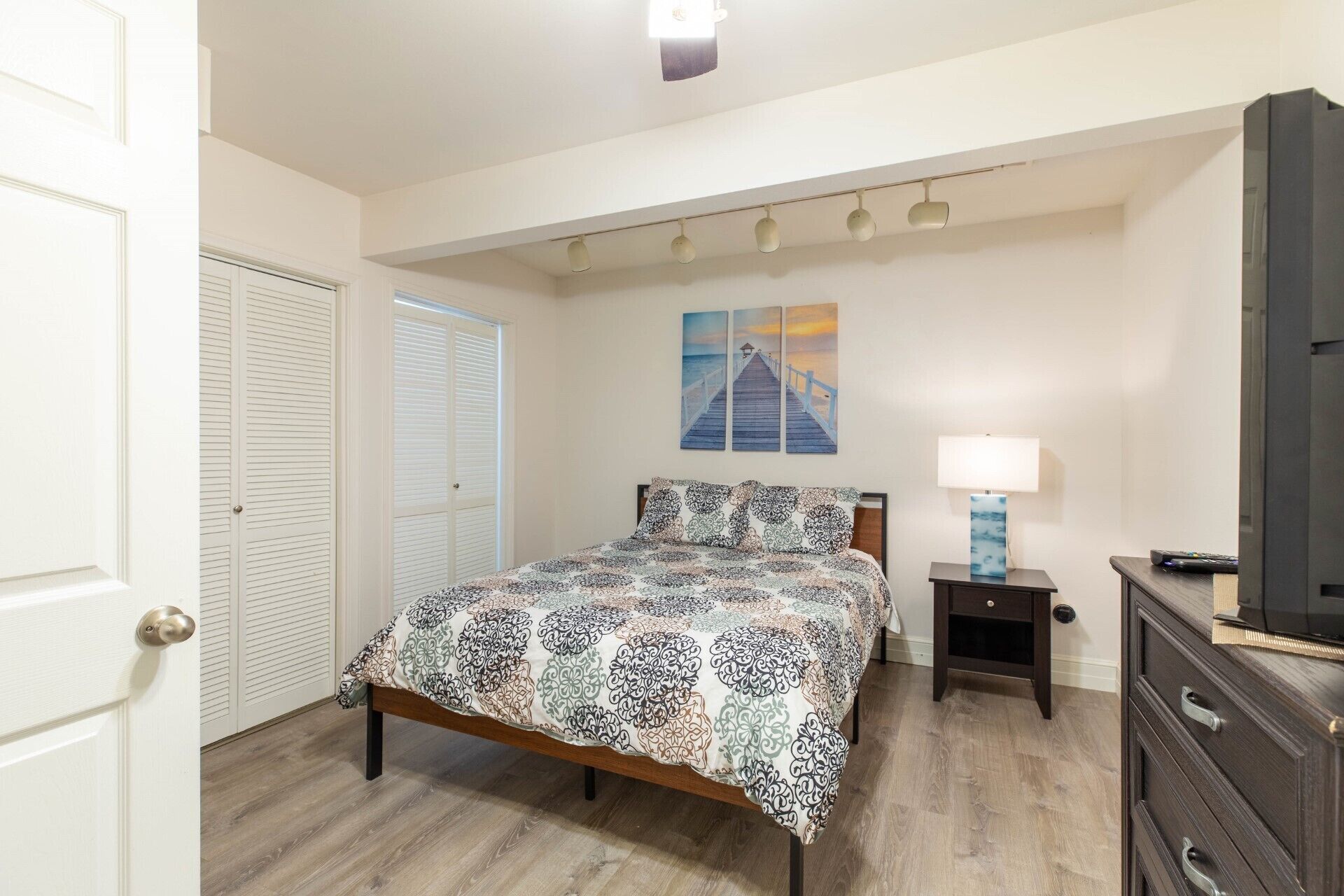 Um quarto com cama, mesa de cabeceira, luminária, cômoda e um quadro na parede.