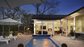 A modern outdoor pool area with a wooden deck, lounge chairs, and a glass-enclosed patio with a fireplace.