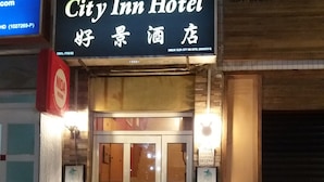 Exterior del Hotel City Inn con puerta de acceso de vidrio, iluminado por luces fijadas en la pared, y una cartelera sobre la entrada.