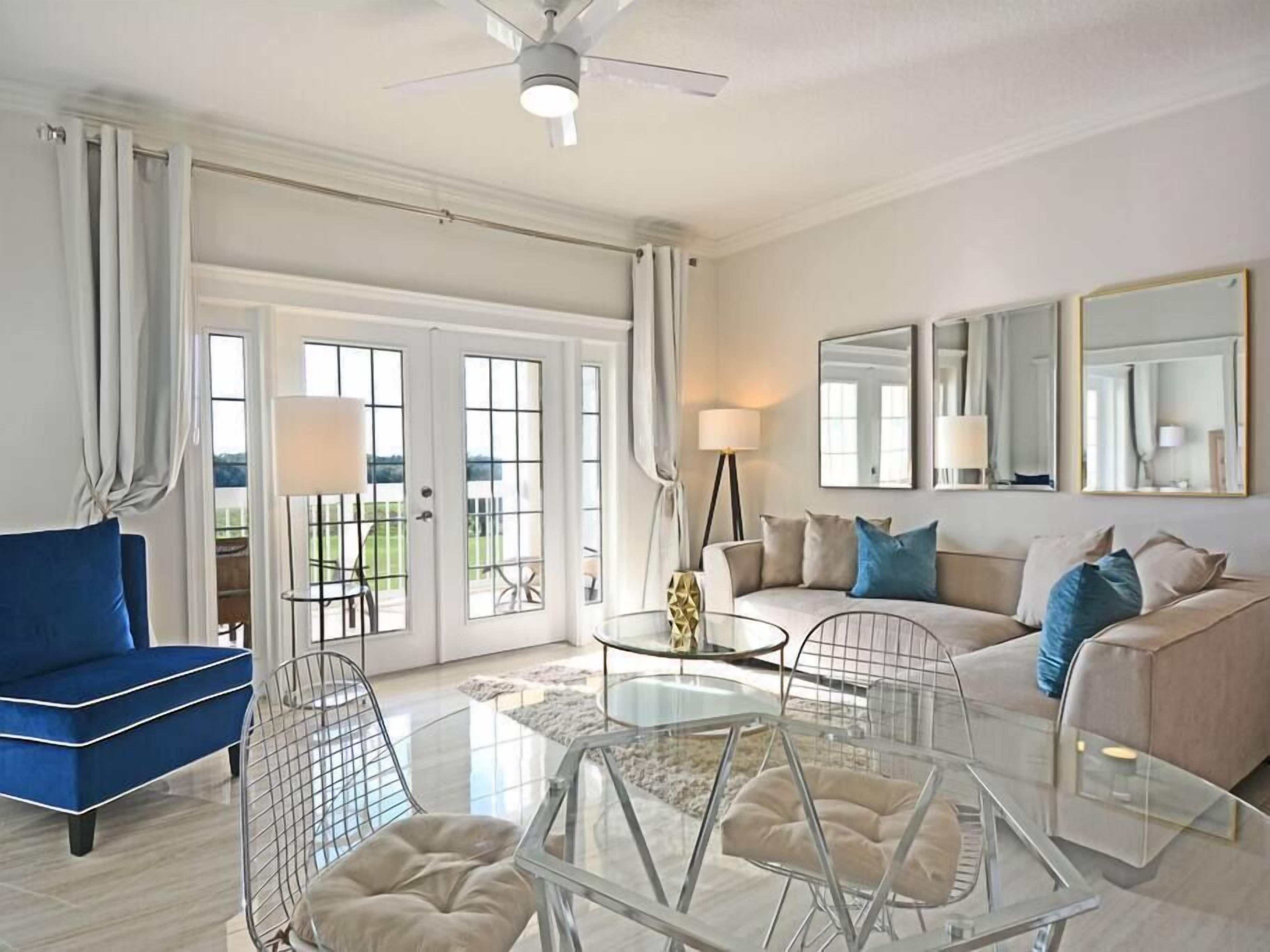 A modern living room with a blue sofa, beige sofa, and a glass coffee table. There are two chairs with cushions, a lamp, and a ceiling fan.