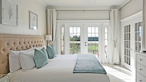 A bedroom with a tufted headboard, a large bed with white and teal bedding, a ceiling fan, and a view through French doors to a balcony. - Style, Luxury & Stunning Views (Reunion)