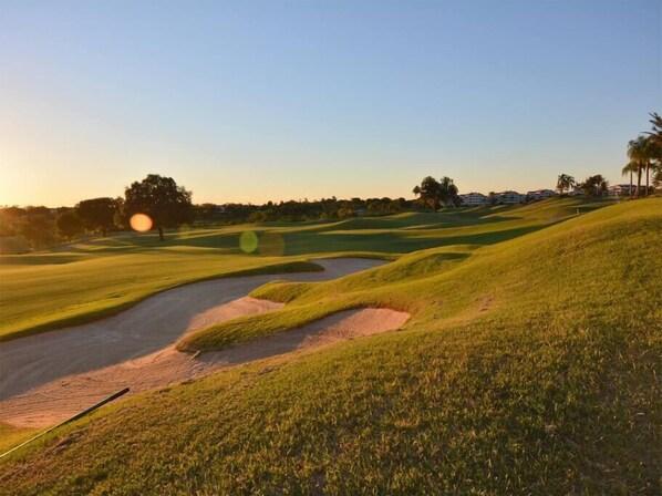 Een golfbaan met zandbunkers, een heldere lucht en vertegaande bomen.