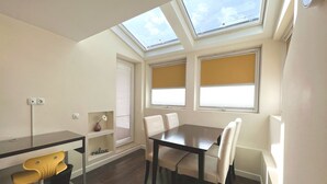 Un comedor con una mesa y sillas, un estante pequeño con un jarrón y una ventana con persianas.