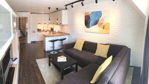 Un salón moderno con un sofá marrón oscuro, dos cojines amarillos, una mesa de centro negra y una revista. Se ve una cocina con armarios blancos y una pared de ladrillo blanco al fondo.