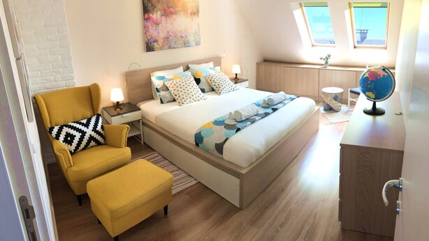 Un dormitorio bien iluminado con una cama, un sillón amarillo, una mesita de noche con una lámpara, un globo terráqueo y un cuadro en la pared.