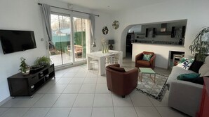 Ein modernes Wohnzimmer mit Flachbildfernseher, einem Essbereich mit weißem Tisch und Stühlen, einem braunen Sessel, einer Couch mit gemusterten Kissen und einer Küchenzeile mit weißer Arbeitsplatte und Schränken.