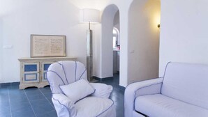 Una sala de estar con un sillón blanco, un sofá, un aparador con un menú enmarcado y una lámpara.