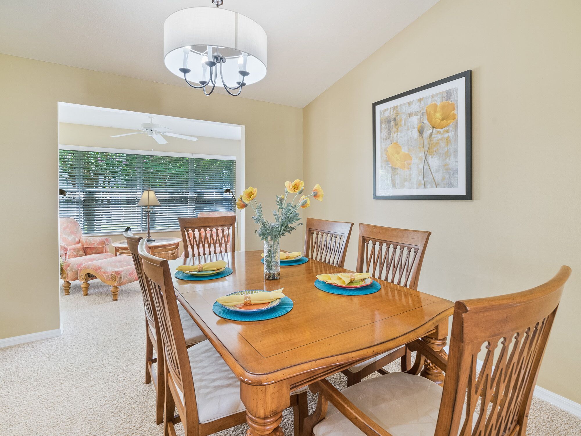 A dining room with a wooden table, chairs, and a chandelier. There is a window with blinds and a framed picture on the wall.