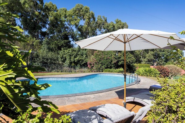 Una piscina con borde de azulejos, rodeada de un jardín con diversas plantas y una sombrilla blanca.
