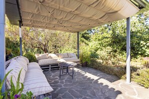 An outdoor patio with a covered seating area, stone flooring, and surrounding greenery.