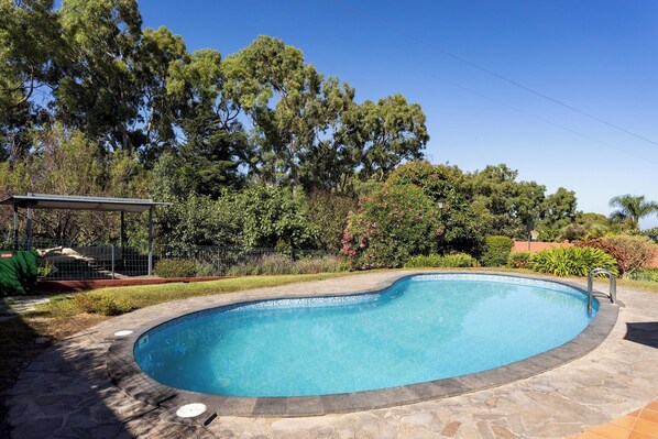 A swimming pool with a stone border, surrounded by trees and a fenced area.