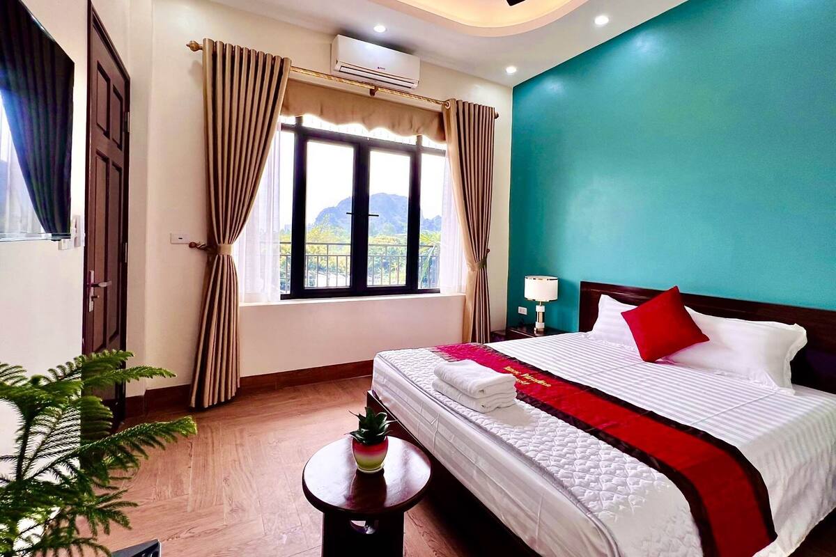 A hotel room with a bed, a nightstand, a lamp, a potted plant, and a window with curtains.