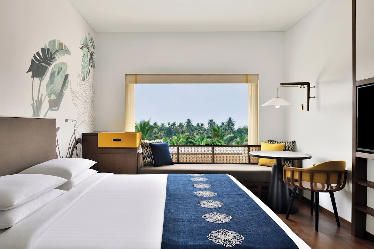 A hotel room with a bed, a desk, a chair, a TV, and a window with a view of greenery.