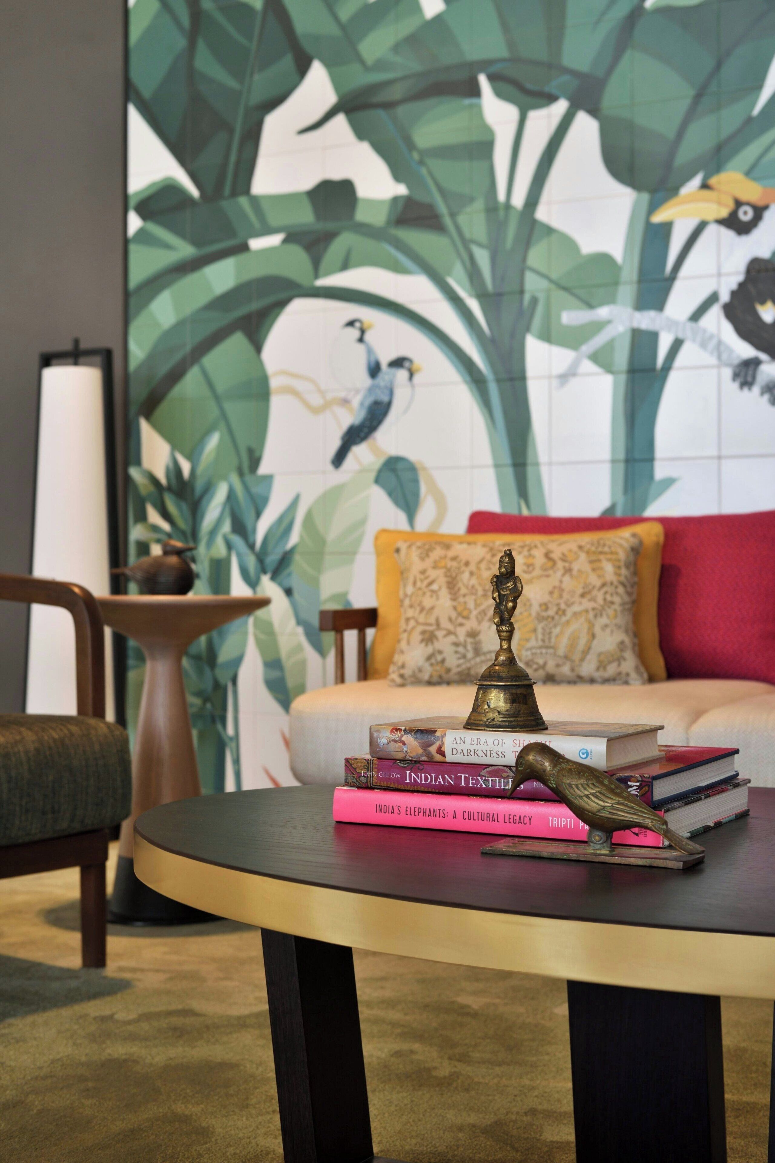 Una mesa redonda con un adorno de latón y libros, un sofá con un cojín rojo, y una pared con un gran mural de aves y follaje.