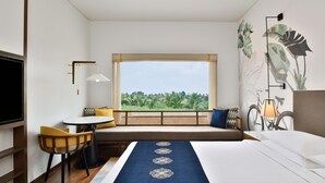 A hotel room with a bed, a seating area with a table and chairs, a TV, a window with a view of greenery, and a bicycle decoration on the wall. - Port Muziris, a Tribute Portfolio Hotel by Marriott, Kochi Airport (Aluva)