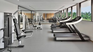 A modern gym with various exercise equipment, large windows, and a view of greenery. - Port Muziris, a Tribute Portfolio Hotel by Marriott, Kochi Airport (Aluva)
