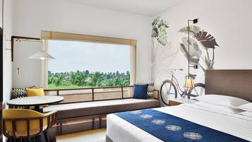 A hotel room with a large window, a bed with a blue bedspread, a seating area with a bicycle, and a dining table with chairs.