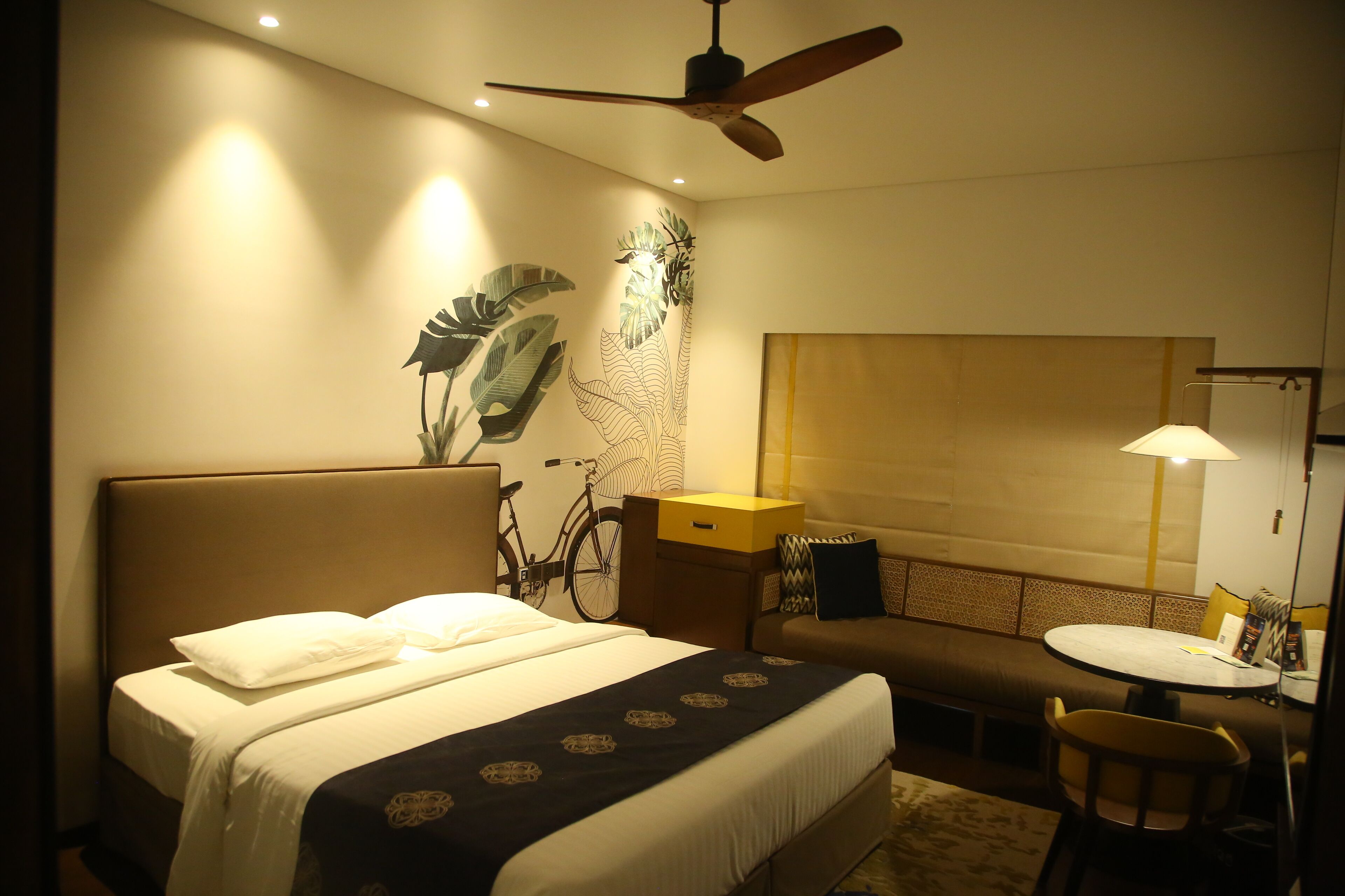Habitación de hotel con cama, una mesa redonda, sofá, bicicleta y pared con diseño de hojas.