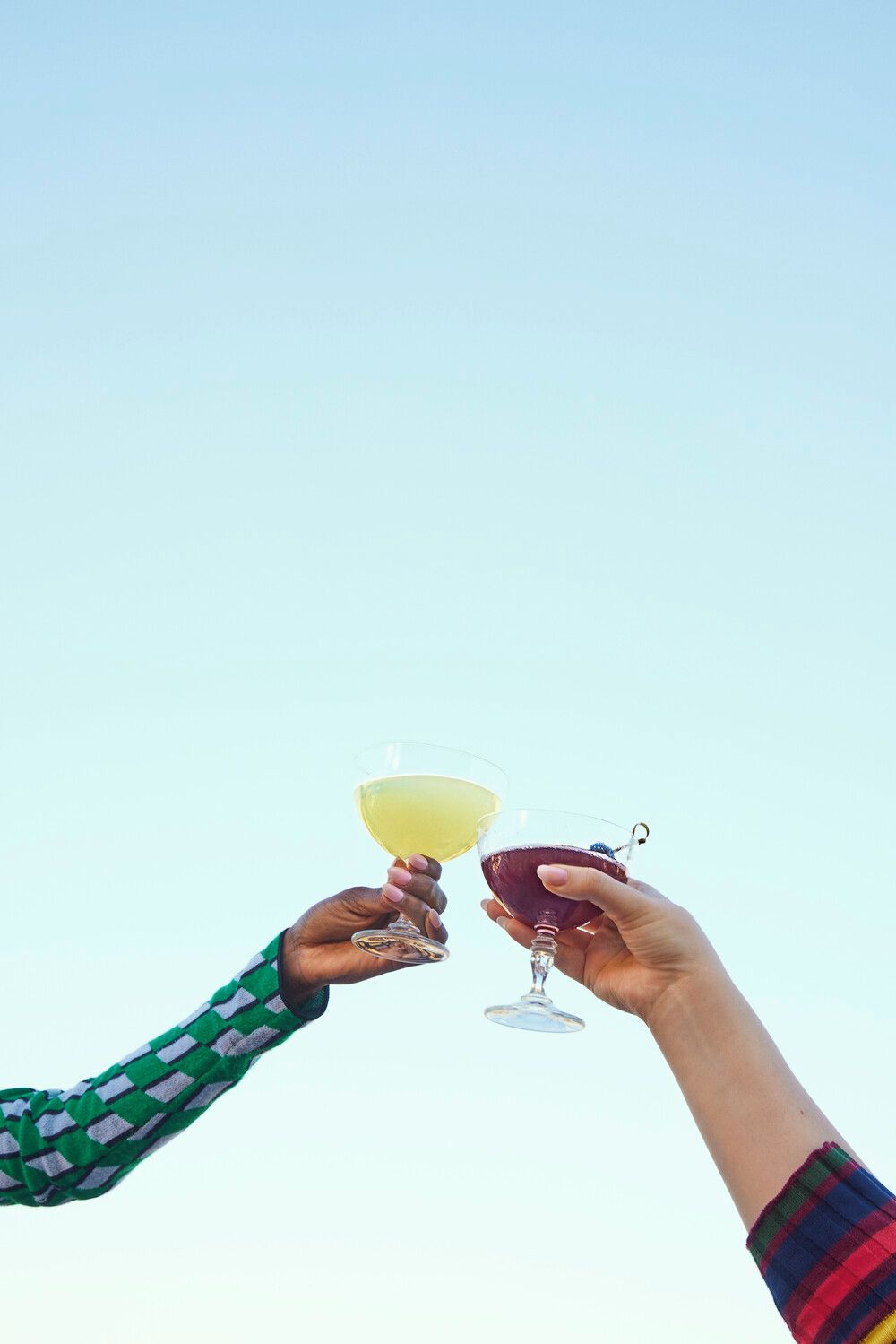 Two hands holding wine glasses against a clear sky.