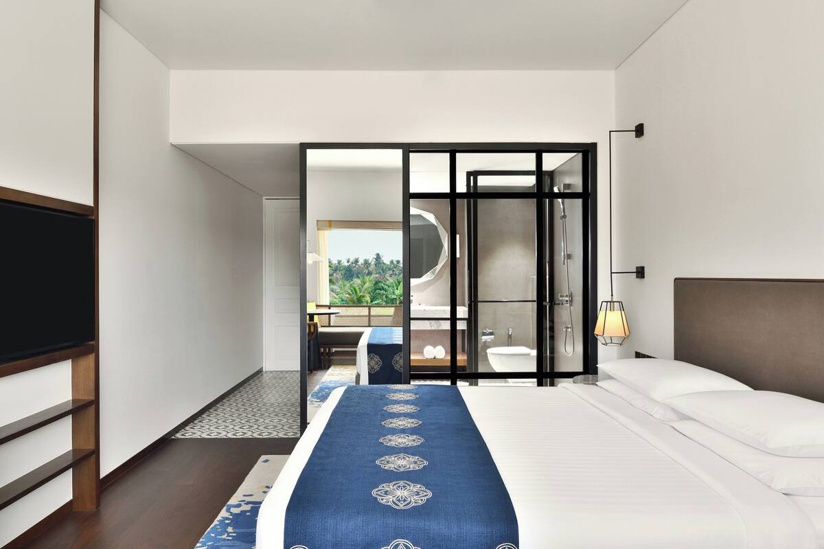 A modern hotel room with a large bed, a TV, a bathroom with a bathtub, and a view of greenery outside.