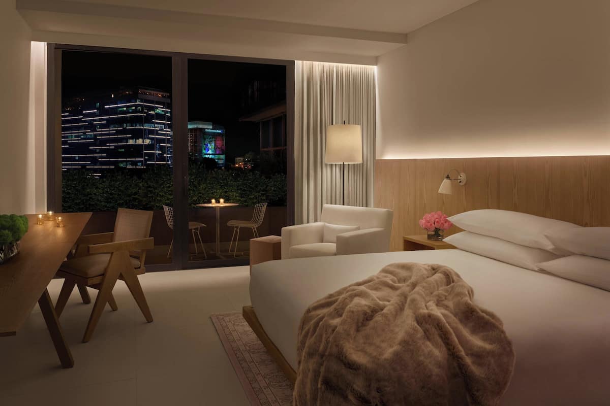 Een moderne hotelkamer met een groot bed, een zithoek met een tafel en stoelen, een nachtkastje met een lamp, en uitzicht op de stad 's nachts.